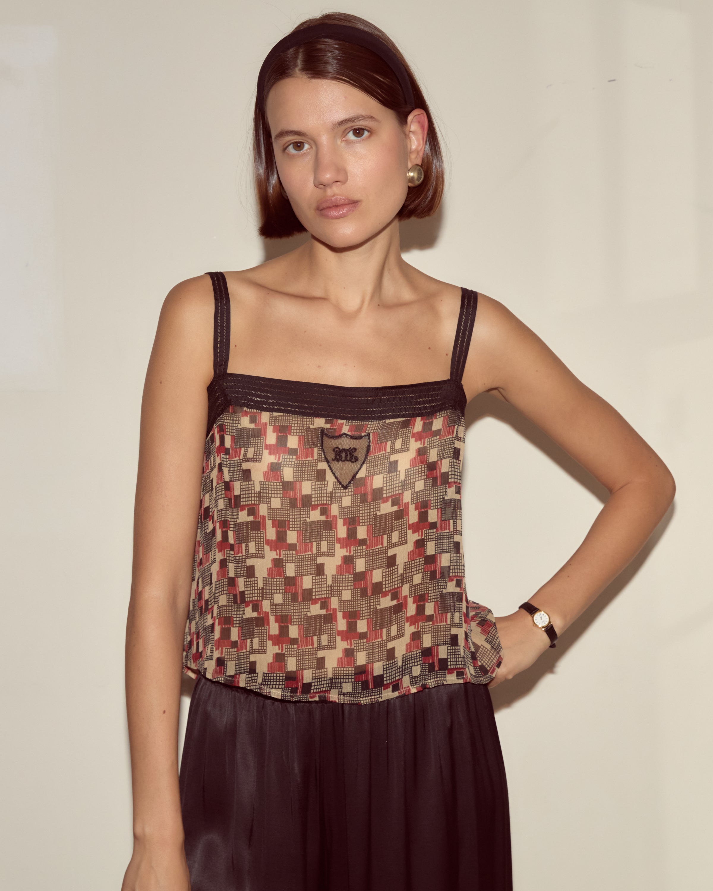 A woman wears the Quadrille Tank, featuring a geometric pattern, paired with a black skirt and stands against a plain light-colored wall.