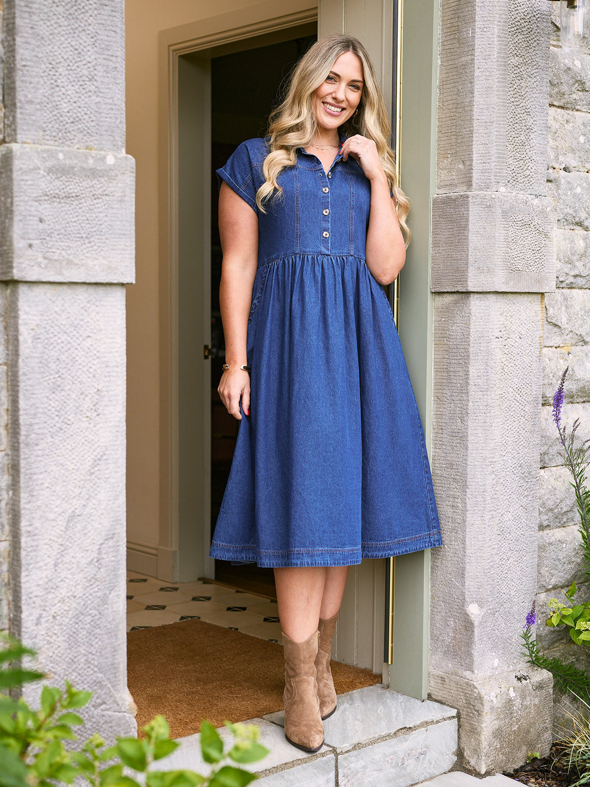Woman in a blue denim dress standing in a doorway of a stone building.