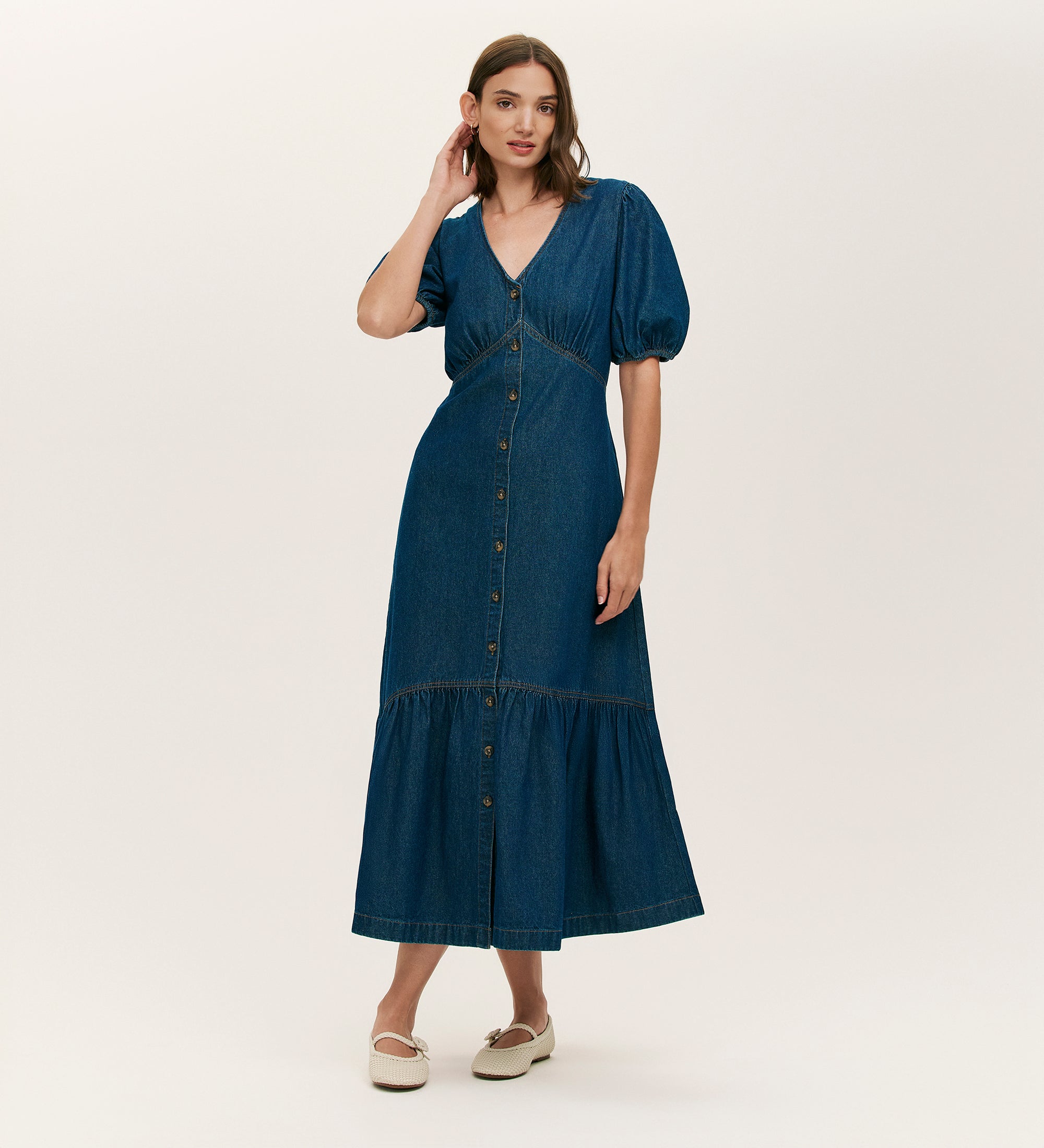 A woman wears the Goldie Indigo Denim Midi Dress, featuring a soft blue denim fabric, short sleeves, V-neckline, button-through front, and midi length. She pairs it with white shoes and stands against an off-white background.