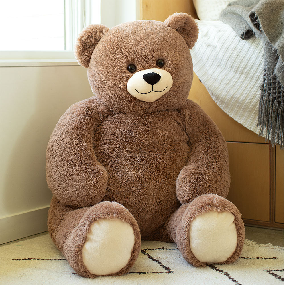 Large plush brown teddy bear with cream-colored paws and a smiling face sitting on a rug near a window and bed.