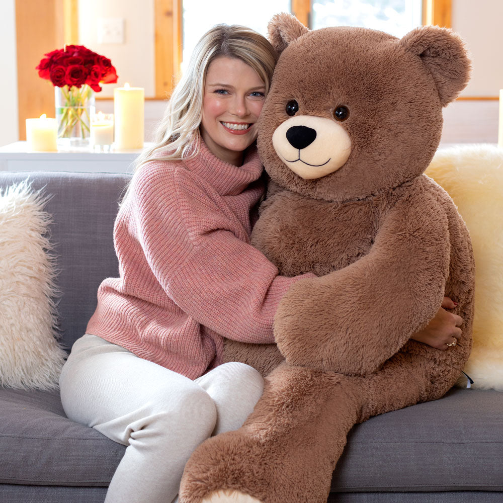 Large plush brown teddy bear with light snout and black eyes, hugged by smiling woman on gray couch.